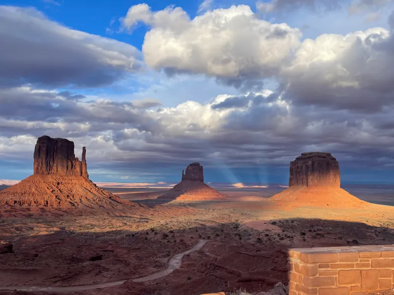 Sun rays illuminating Monument Valley rock formations under cloudy sky.
