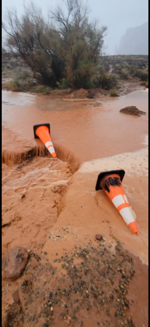 Two traffic cones half-submerged in muddy floodwater on a dirt road.